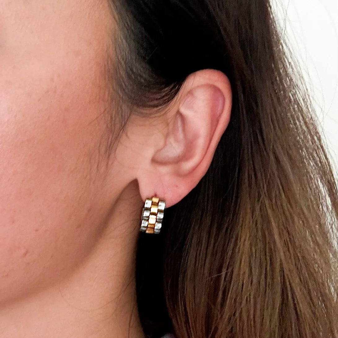 A close-up side profile of a woman's ear wearing a small checkered hoop earring in silver and gold-tone metal.