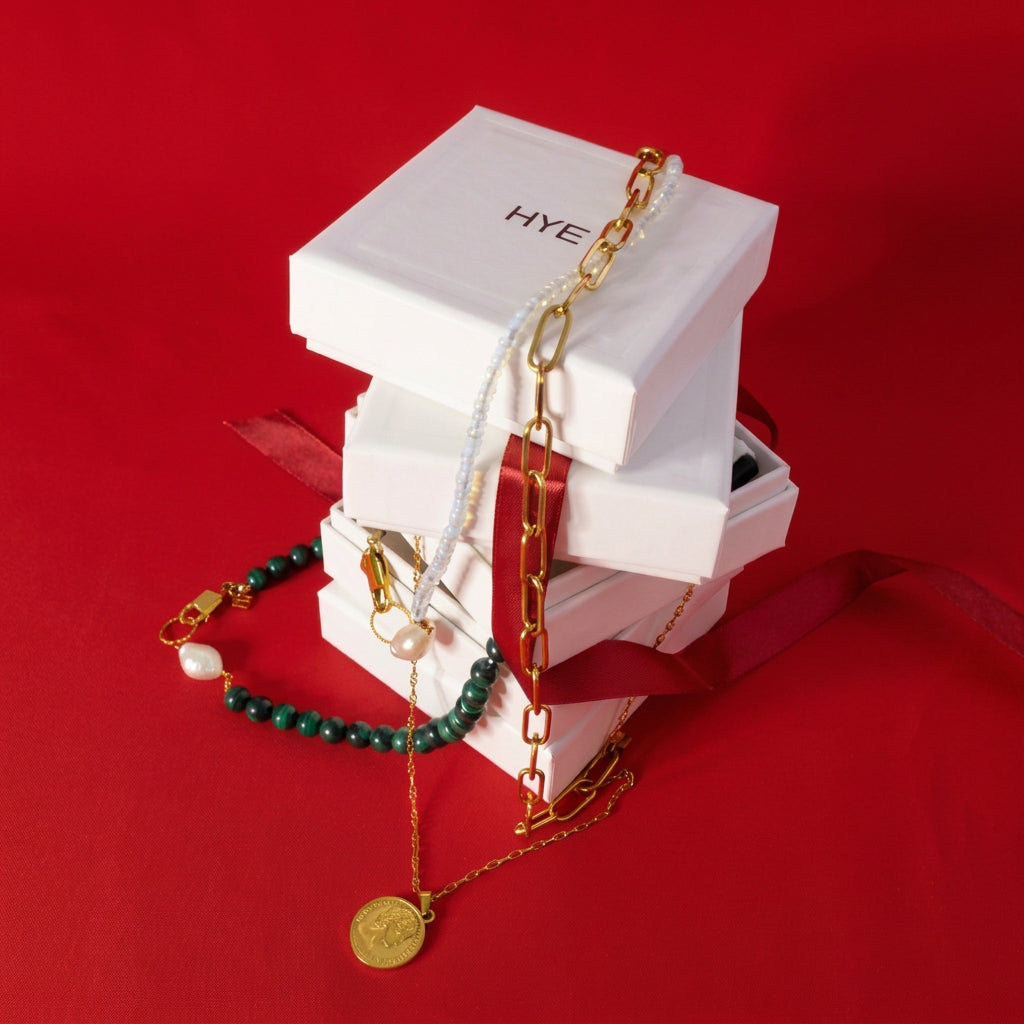 A stack of white jewelry boxes branded HYE, styled with multiple gold-tone necklaces and bracelets, including a green beaded bracelet and a gold coin pendant, set against a vibrant red textured fabric background.