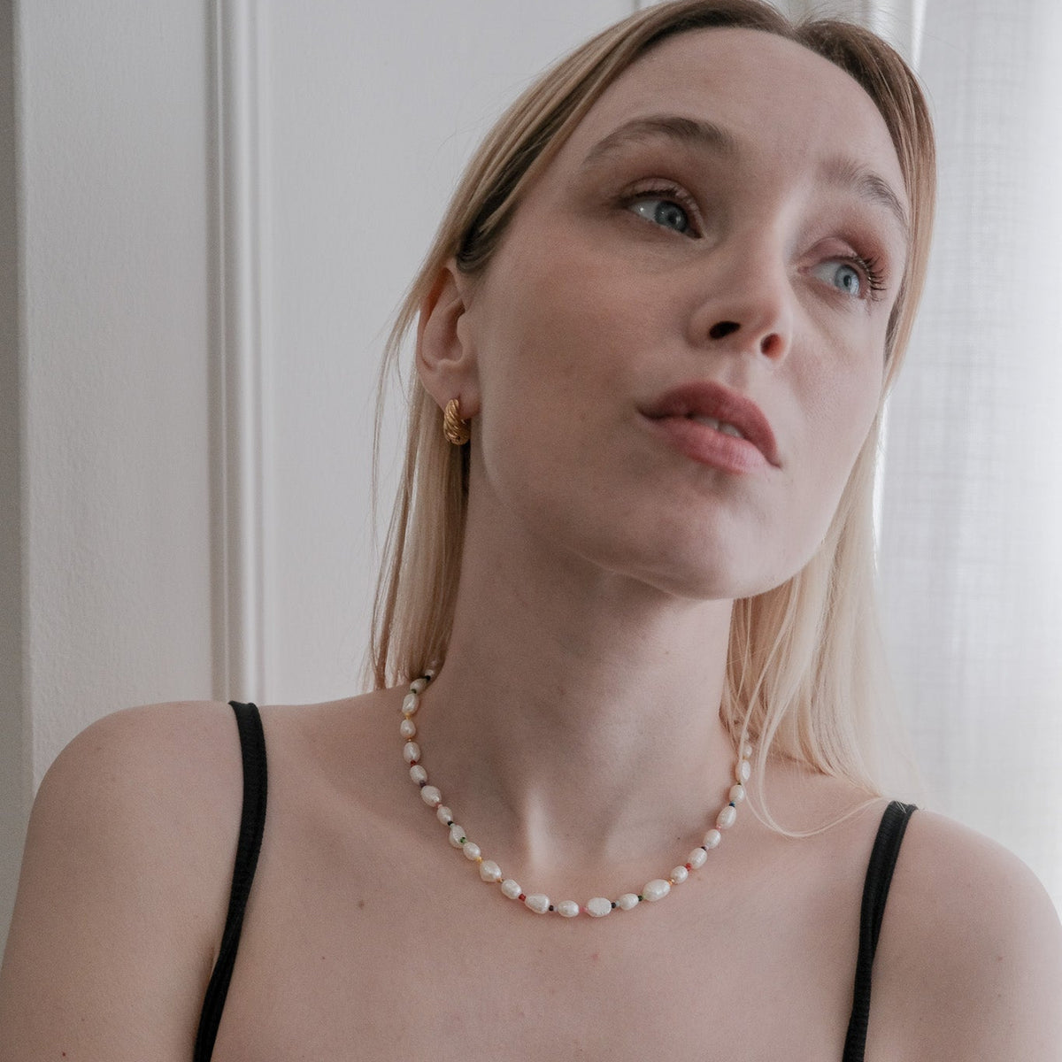 A blonde woman wearing a black ribbed camisole top and a white pearl necklace, looking upward and to the side in a softly lit indoor setting.
