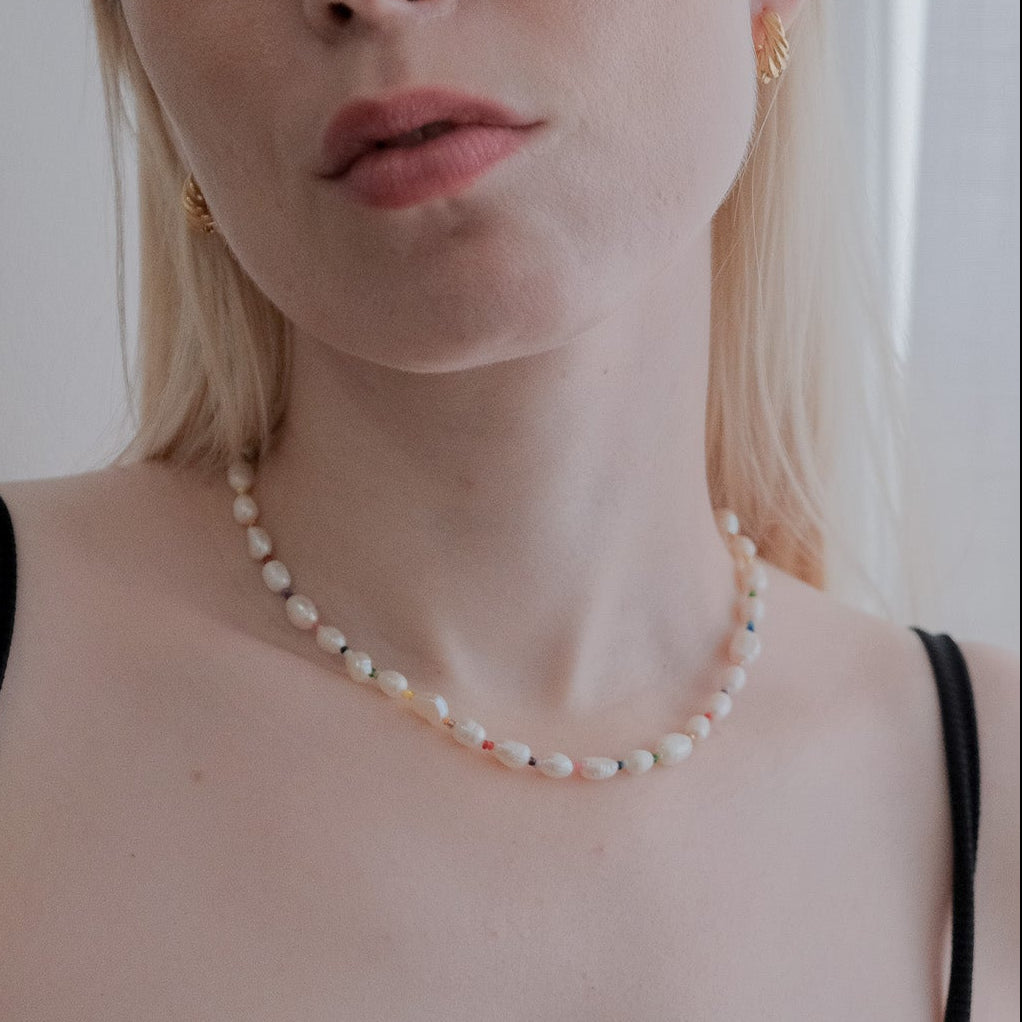 A close-up shot of a woman with blonde hair wearing a delicate white pearl necklace and a black spaghetti-strap top, set against a soft, light-colored indoor background.