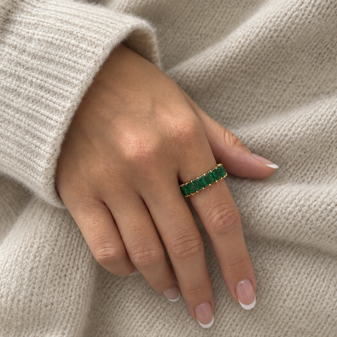 A close-up of a person's hand wearing a gold ring with a row of green rectangular stones, resting against a cozy beige knit sweater.
