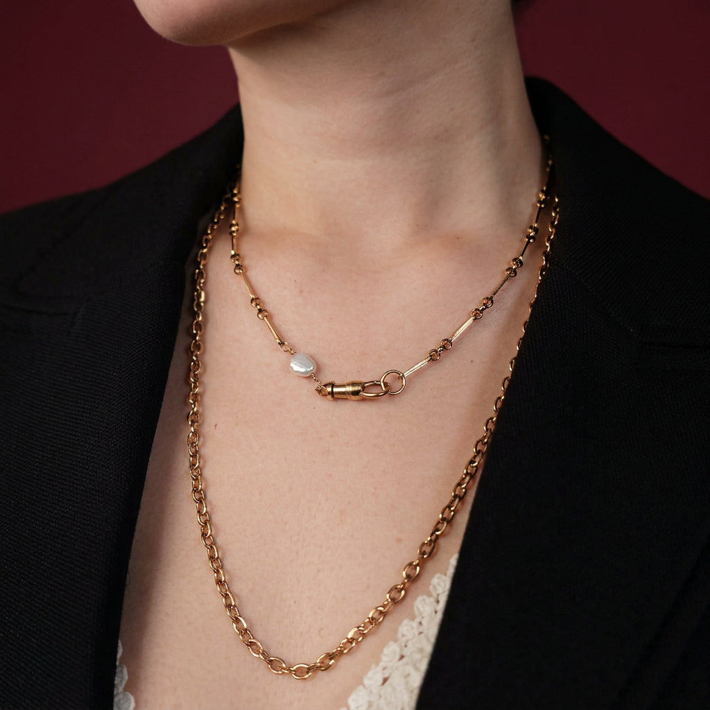 A close-up of a person wearing two layered gold-tone necklaces. The shorter chain features a small white teardrop-shaped stone pendant, while the longer chain has a textured link design.