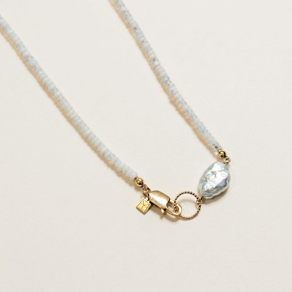A delicate necklace featuring a white beaded strand, a gold lobster clasp, a small gold charm, a silver-tone ring, and a polished silver baroque stone, displayed on a textured white surface.
